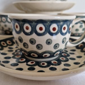 May include: A set of ceramic teacups and saucers. The cups and saucers are white with a blue and red pattern. The cups have a handle and the saucers are round. The pattern consists of blue circles with red dots in the center.