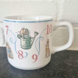May include: White ceramic mug with a blue rim and handle. The mug features illustrations of a rabbit, watering can, and numbers 7, 8, 9, and 10 in red. A whimsical design for a kitchen or home decor.