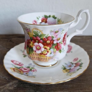 May include: A white teacup and saucer with a floral pattern. The cup and saucer feature a basket of pink, red, and yellow flowers with green leaves. The rim of the saucer is gold.