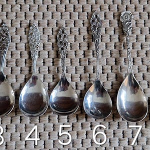 Könnte beinhalten: Eine Sammlung von zehn antiken Silberlöffeln, jeder mit einem einzigartigen Design. Die Löffel variieren in Form und Stil, mit verzierten Griffen und verschiedenen Schalenformen. Sie sind in einer Reihe auf einer strukturierten Oberfläche angeordnet, nummeriert von 1 bis 10.