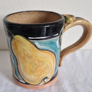 May include: A ceramic mug with a hand-painted design of a yellow pear on a blue and black background. The mug has a brown handle.