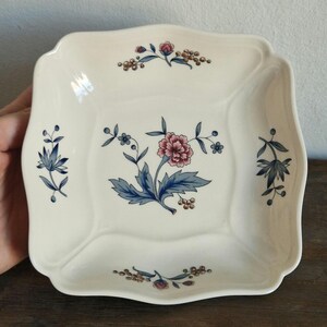 Puede incluir: Un plato cuadrado de cerámica blanca con un borde festoneado. El plato presenta un diseño floral en tonos azules, rosas y marrones.
