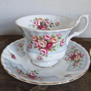 May include: A white teacup and saucer set with a floral rose pattern in shades of pink and peach. The cup and saucer have a light blue border and gold trim. The teacup is on a wooden surface.
