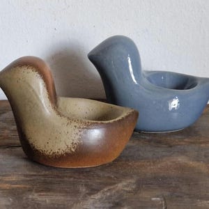 May include: Two ceramic bird-shaped egg cups. One is brown with a speckled glaze, the other is a light blue.