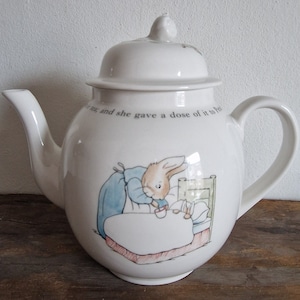 May include: White ceramic teapot with a lid, featuring a Peter Rabbit illustration. The teapot has a curved spout and handle, with text around the top. The illustration shows Peter Rabbit in bed. A charming addition to any kitchen.