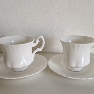 May include: Two white teacups and saucers with a delicate, scalloped design. The cups have a classic shape with a rounded base and a flared rim.