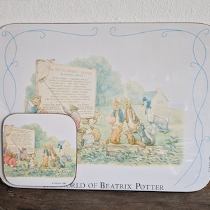 Puede incluir: Un mantel individual rectangular blanco y azul con un borde de espirales azules. El mantel individual presenta una imagen de los personajes de Peter Rabbit de Beatrix Potter reunidos alrededor de un cartel que dice "The Peter Rabbit Books by Beatrix Potter". El cartel enumera los títulos de los libros. El mantel individual tiene el texto "The World of Beatrix Potter" en la parte inferior.