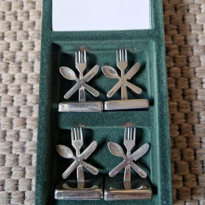 Op de afbeelding: Set van vier zilverkleurige metalen placecardhouders. Elke houder heeft een ontwerp van een vork, mes en lepel in een kruispatroon.
