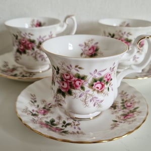May include: Three white teacups and saucers with a floral pattern of pink roses and green leaves. The cups have a gold rim.