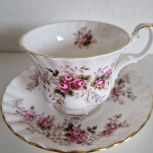 May include: A white teacup and saucer set with a floral pattern of pink roses and green leaves. The set has a gold trim around the edges.