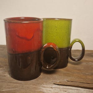 Può includere: Due tazze in ceramica con smalto marrone, rosso e verde. Le tazze hanno una forma arrotondata e un manico.