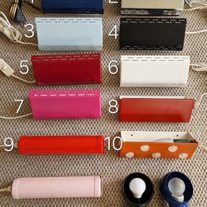 Op de afbeelding: Een verzameling vintage elektrische lampen in verschillende kleuren, waaronder blauw, zilver, lichtblauw, zwart, rood, wit, roze en oranje. De lampen hebben rechthoekige en cilindrische vormen, met zichtbare snoeren en stekkers. Ook inbegrepen zijn twee kleine lampen met gloeilampen.