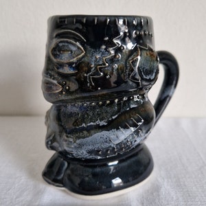 Op de afbeelding: Een zwarte keramische tiki-mok met een gedetailleerd gezicht en een handvat. De mok heeft een gespikkelde glazuur en een witte basis.