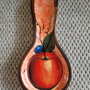 Peut inclure: Repose-cuillère en céramique avec un motif de pomme peint à la main. Le repose-cuillère présente une pomme rouge avec une feuille verte et une baie bleue. La bordure est marron et le fond est orange.