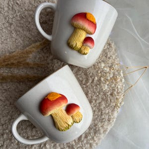 以下が含まれることがあります： 白の陶器マグカップ2つ。それぞれに3Dのキノコのデザインが施されています。キノコは赤と黄色の傘と黄色の茎が特徴です。マグカップはカーブしたハンドルが付いており、ニュートラルな質感の背景に置かれています。