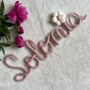 Puede incluir: Un cordón tejido de color rosa que forma el nombre Selma en cursiva, decorado con una flor de ganchillo blanca en la parte superior derecha. Tres peonías rosas vibrantes con hojas verdes se sitúan a la izquierda del nombre tejido.