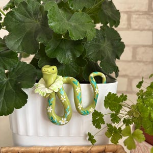 May include: A light green snake figurine with a white ruffled collar, featuring hand-painted white flowers and teal leaves along its body, designed to hook onto the edge of a white planter pot with vertical ridges.