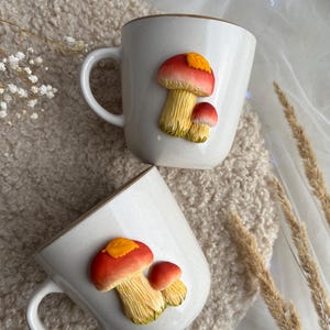 以下が含まれることがあります： キノコのデザインが施された2つの白いセラミックマグ。キノコは、黄色いアクセントと黄色の茎が付いた赤い帽子をしています。マグカップはまだらの仕上げと茶色の縁があります。マグカップはクリーム色のテクスチャード加工された表面にあります。