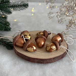 May include: A collection of five decorative acorns featuring metallic copper-toned shells and textured brown caps. Each acorn is accented with patches of gold leaf and includes a thin white string loop attached to the top for hanging purposes.