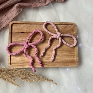May include: Two pink knitted bows with long, dangling tails sit on a wooden tray. The bows are a soft, pastel pink colour, and the texture of the knit is visible. The background includes a pink fabric and dried plants.
