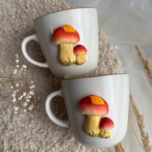 以下が含まれることがあります： キノコの模様が入った白いセラミックマグカップ2つ。各マグカップには、赤い帽子と黄色のひだのある大きなキノコと、小さなキノコが描かれています。マグカップには茶色の縁とハンドルが付いています。