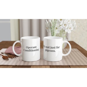 Puede incluir: Dos tazas de cerámica blanca con texto negro. La taza de la izquierda dice "Condicionamiento operante". La taza de la derecha dice "No es solo para palomas."