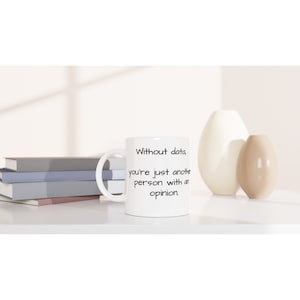 Peut inclure: Mug en céramique blanche avec le texte "Without data, you're just another person with an opinion." La tasse a une anse incurvée et est posée sur une surface blanche. Des vases décoratifs et des livres sont en arrière-plan.