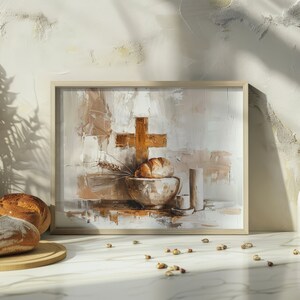 Puede incluir: Una pintura abstracta en un marco de madera representa una cruz dorada, pan y un cuenco. Dos hogazas de pan están sobre una tabla de cortar de madera. Una botella blanca y un jarrón con utensilios de madera están a la derecha. La escena está sobre una superficie de mármol.