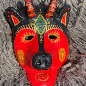 May include: A hand-painted mask with a black base, red face, and gold accents. The mask features a stylized animal design with horns, ear-like shapes, and a diamond-shaped emblem. The mask has eye holes and a nose, and is decorated with orange and yellow details.