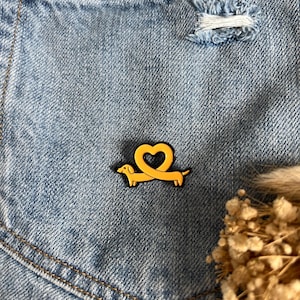 Könnte beinhalten: Gelber Dackel-Ansteckpin mit herzförmigem Schwanz, vor hellblauem Jeanshintergrund. Der Pin ist schwarz umrandet und hat ein glänzendes Finish. Der Jeansstoff hat sichtbare Nähte und ein Used-Detail.
