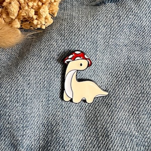 Könnte beinhalten: Ein skurriler Emaille-Pin mit einem hellgelben Dinosaurier mit rotem und weißem Pilzhut. Der Dinosaurier hat einen weißen Bauch und einen schwarzen Umriss. Der Pin ist vor einem Jeanshintergrund platziert.