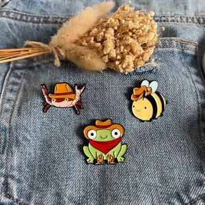 Könnte beinhalten: Drei Emaille-Anstecknadeln mit Cartoon-Tieren, die Cowboyhüte tragen. Ein Krebs, eine Biene und ein Frosch sind in leuchtenden Farben dargestellt. Die Anstecknadeln sind vor einem Jeansstoff-Hintergrund platziert, mit einem kleinen Strauß Trockenblumen.