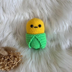 Könnte beinhalten: Ein handgefertigtes gehäkeltes Maiskolben-Plüschtier. Der Mais hat einen gelben Kopf mit schwarzen Augen und einem kleinen Mund. Die grüne Hülle umschließt den gelben Mais. Das Plüschtier befindet sich vor einem weißen, flauschigen Hintergrund.