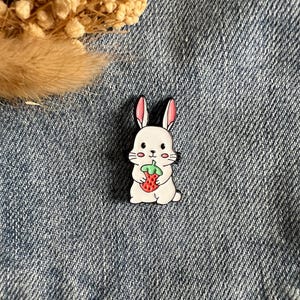 Könnte beinhalten: Weißer Hasen-Ansteckpin mit rosa Ohren und schwarzer Umrandung, der eine rote Erdbeere mit grünen Blättern hält. Der Pin befindet sich vor einem blauen Jeans-Hintergrund.