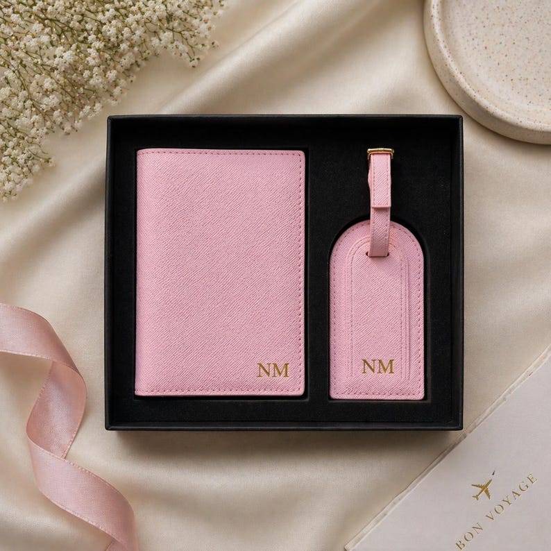 May include: A light pink passport holder and a matching luggage tag with a strap, both featuring the gold embossed initials NM, presented in a black rectangular gift box with a black interior lining and a textured surface finish.