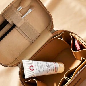 May include: A tan cosmetic travel case with a zip closure, featuring an interior with elastic brush holders and multiple storage compartments. Inside, a tube of First Aid Beauty Pure Skin Face Cleanser is visible, along with two makeup brushes.