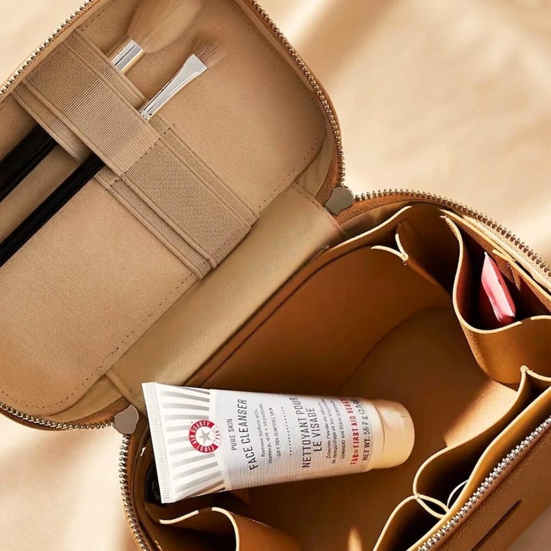 May include: A tan cosmetic travel case with a zip closure, featuring an interior with elastic brush holders and multiple storage compartments. Inside, a tube of First Aid Beauty Pure Skin Face Cleanser is visible, along with two makeup brushes.