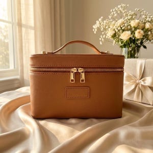 May include: A rectangular tan vanity case with a top handle and gold-toned hardware. The case features a double zip closure across the top and a small rectangular patch on the front centre. The exterior has a textured finish and structured design.
