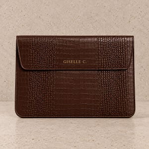 May include: A dark brown rectangular laptop sleeve featuring a textured crocodile skin pattern. The front flap is embossed with the text GISELLE C. in gold lettering. The sleeve has a structured design with a fold-over closure and rounded corners.