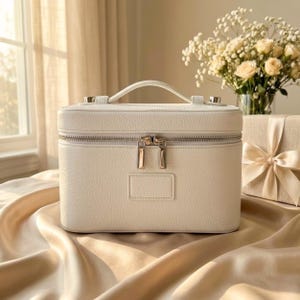 May include: A rectangular white vanity case with a textured finish, featuring a top handle, a silver-toned double zipper closure, and a small rectangular patch on the front. The case has a structured design with rounded corners and metallic hardware.