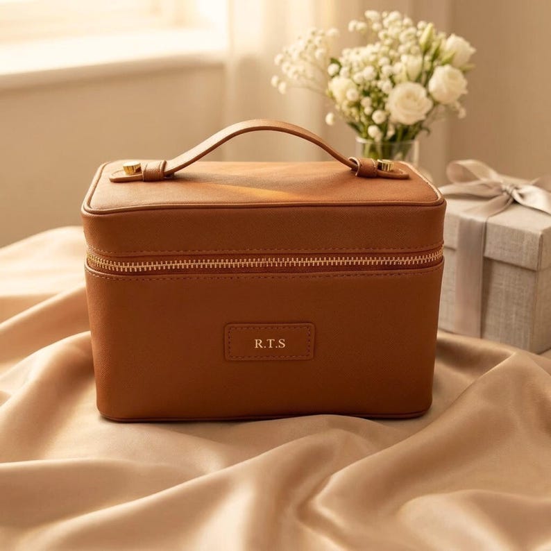 May include: A rectangular tan vanity case with a top handle and a gold-toned zip closure. The front features a small rectangular patch embossed with the letters R.T.S. The case has a smooth, structured exterior and a clean, minimalist design.