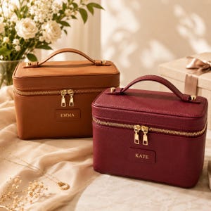 May include: Two rectangular vanity cases with top handles and gold-tone zips. One case is tan with the name EMMA embossed on a front patch, and the other is deep red with the name KATE embossed on a front patch. Both feature a structured design.