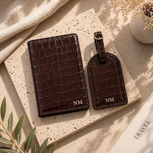 May include: A dark brown passport holder and a matching luggage tag, both featuring a crocodile-embossed texture and the gold-embossed initials NM. The passport holder is rectangular, and the luggage tag has a gold-toned metal buckle.