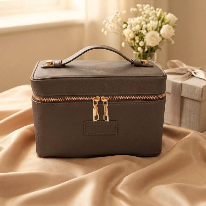 May include: A rectangular dark grey cosmetic case with a top handle and a gold-toned zip closure featuring two matching metal pulls. The bag has a smooth, solid-coloured exterior with a small rectangular patch centred on the front panel.