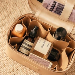 May include: A beige leather cosmetic bag with a zip closure and internal dividers. The bag contains various skincare and fragrance bottles, including Aesop and Byredo Young Rose products, along with a small rectangular box of matches inside.