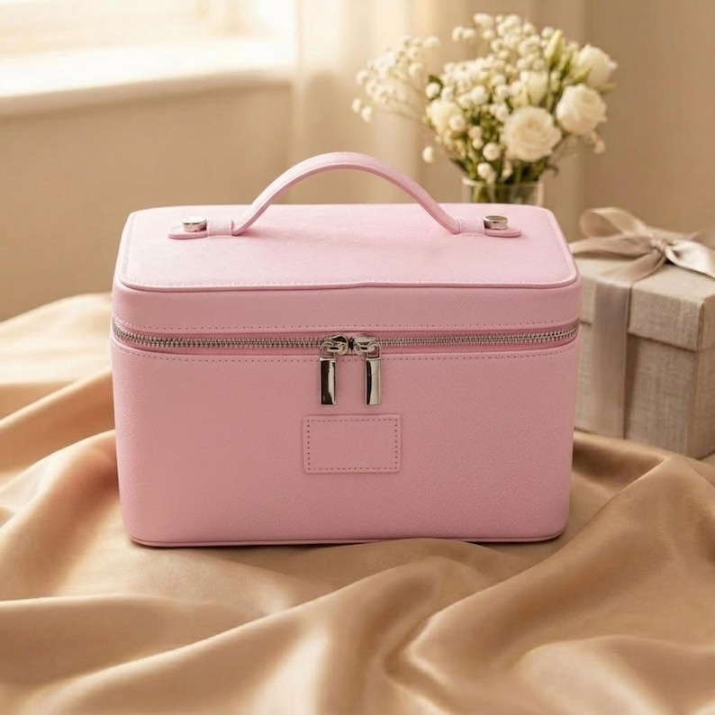 May include: A rectangular light pink vanity case with a top handle, a silver-toned double zip closure, and a small rectangular patch on the front. The case features a textured exterior finish and is constructed with a structured, boxy silhouette.