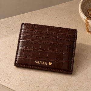 May include: A dark brown rectangular card holder with a crocodile-embossed texture. The front features three horizontal card slots and the gold-coloured text SARAH followed by a small heart symbol printed near the bottom edge of the accessory.