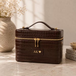May include: A dark brown rectangular vanity case with a crocodile-embossed texture, featuring a top handle, gold-toned metal zippers, and a rectangular patch on the front engraved with the letters AK and a heart symbol in a gold-toned font.