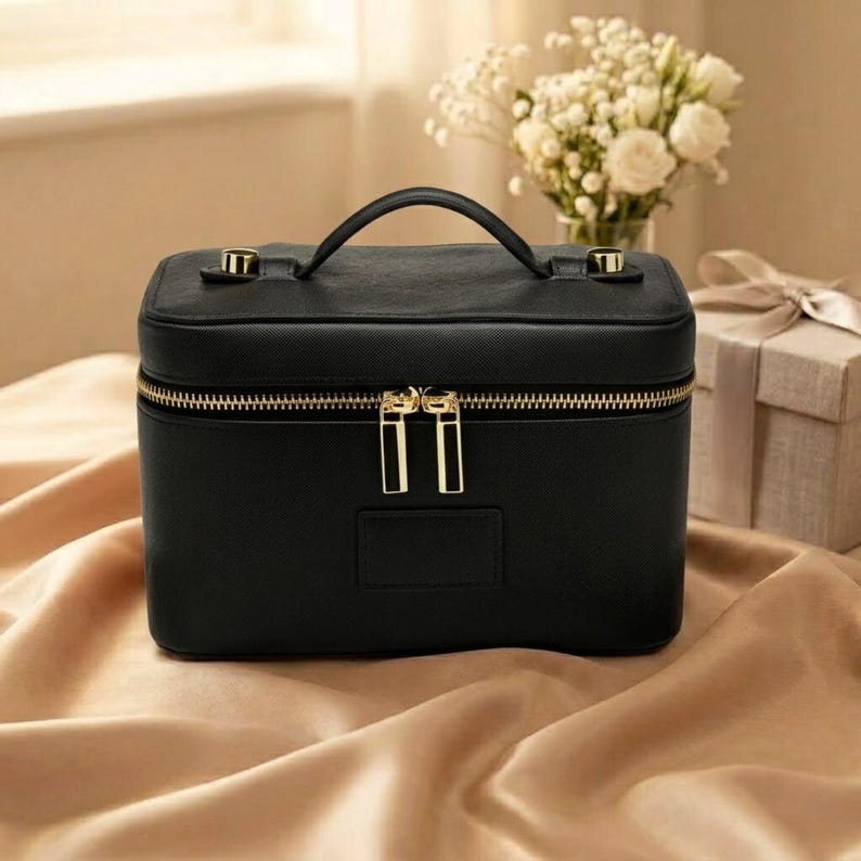 May include: A black rectangular vanity case with a top handle and gold-toned hardware. The case features a double zip closure with two rectangular gold zip pulls and a smooth, solid black exterior finish with a structured, boxy silhouette.