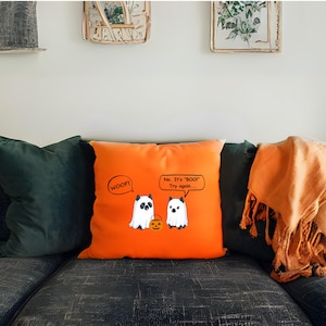 May include: Orange square pillow with a Halloween-themed design. The pillow features two cartoon ghosts, one saying "WOOF!" and the other saying "No. It's 'BOO!' Try again..." A small jack-o'-lantern is between them.